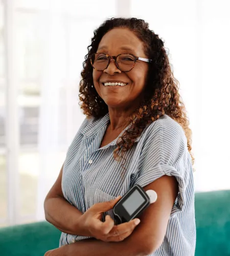 Smiling woman using a glucose meter on her arm indoors near a green sofa with natural light.
