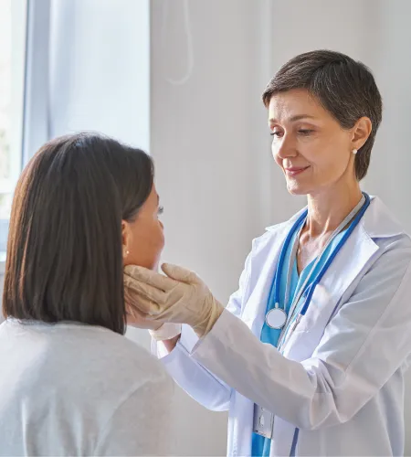 Female doctor in white coat examining patient's throat in medical office with natural light window.