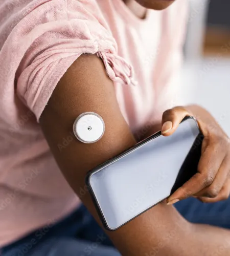 Person wearing a glucose monitor on arm while holding a smartphone, in casual pink shirt and jeans.