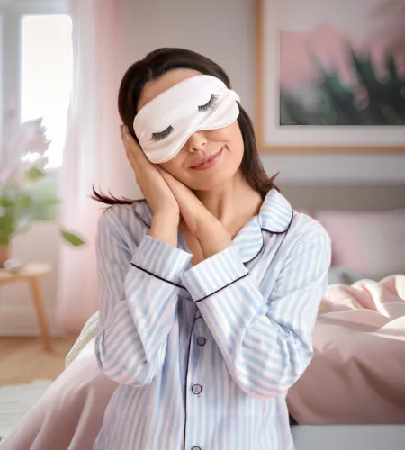 Woman in striped pajamas wearing sleep mask and resting hands on her cheek in cozy bedroom setting