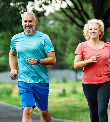 Middle-aged man and woman jogging outdoors in athletic wear with greenery in the background, smiling and exercising.