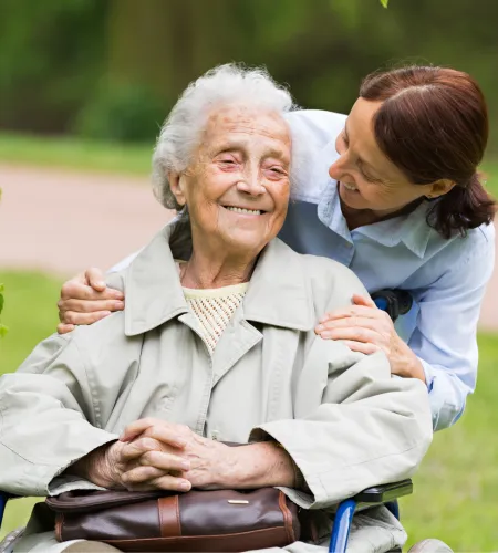 Smiling elderly woman in wheelchair outdoors with caregiver standing behind, showing support and affection.