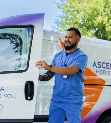 Smiling healthcare worker in blue scrubs opening medical service van door outdoors on a sunny day