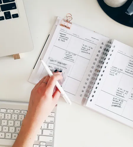 Person writing in a weekly planner beside a keyboard and laptop on a white desk workspace.