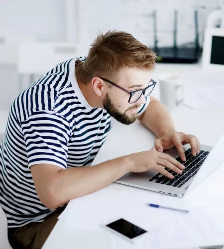 Young man in striped shirt and glasses typing on laptop at a white desk in a bright room.