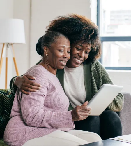 Two women smiling while looking at a tablet together in a cozy living room setting.