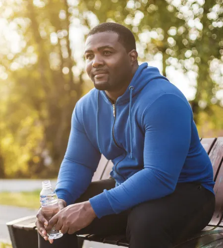 Man in blue hoodie sitting on a bench outdoors holding a water bottle with a thoughtful expression