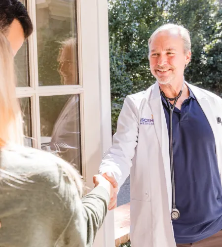 Smiling doctor in white coat shaking hands with patients at home near glass door in sunlight