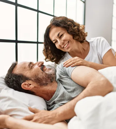 Smiling middle-aged couple relaxing and sharing a tender moment in bed by a large window with natural light.