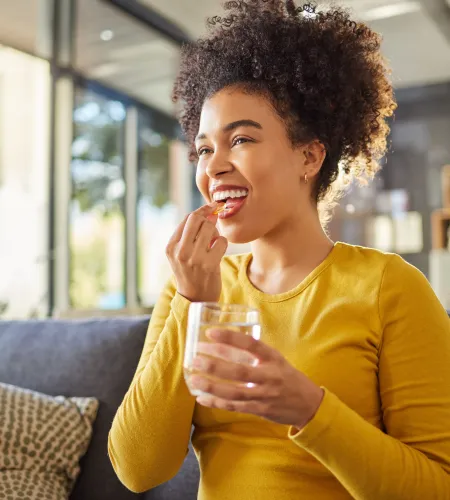 Smiling woman in yellow sweater taking a pill with a glass of water while sitting on a couch at home.