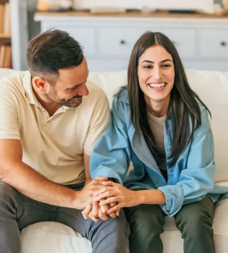 Couple sitting on a couch smiling and holding hands in a cozy living room setting.