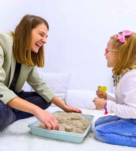Woman and young girl play with kinetic sand in a tray, smiling and sitting on a soft white surface indoors.