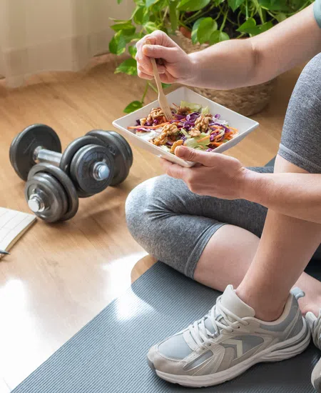 Person in workout clothes sitting on yoga mat eating healthy salad next to dumbbells and meal plan notebook