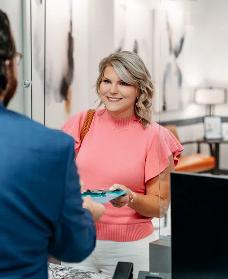 Smiling woman in pink top handing a clipboard to a man in a blue suit at a modern office reception desk.