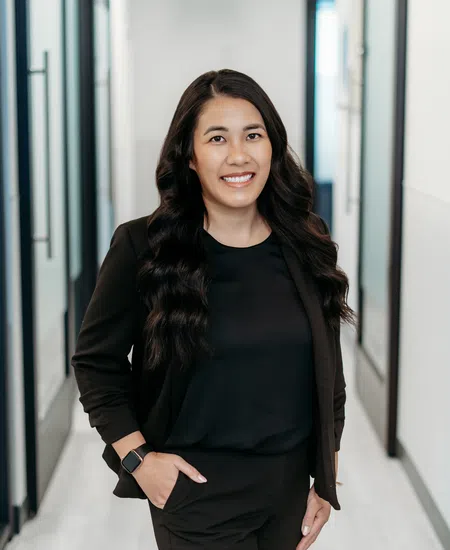Smiling woman with long wavy hair in a black suit standing confidently in a modern office hallway.