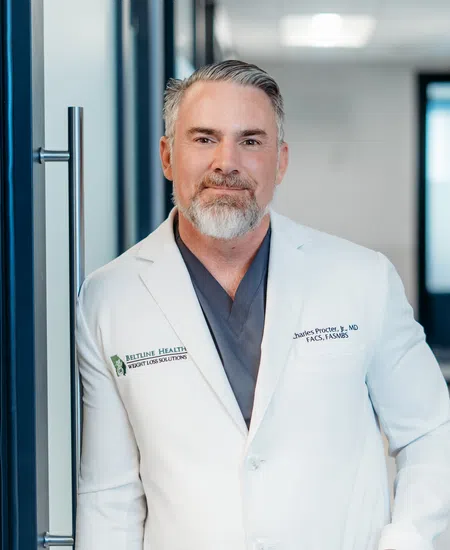 Confident male doctor with grey hair and beard wearing a white coat standing in a medical office hallway