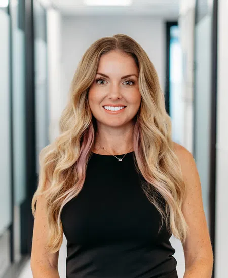 Smiling woman with long blonde hair wearing a black sleeveless dress standing in a bright modern hallway.
