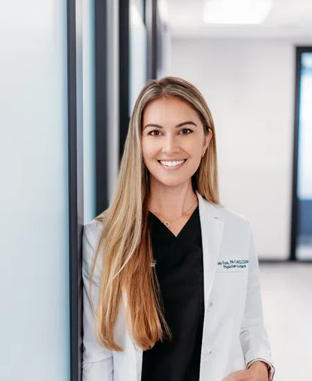 Smiling female doctor with long blonde hair wearing a white coat and black scrubs in a modern clinic corridor.