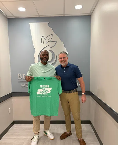 Two men standing indoors holding a green shirt that says Missing 30 Pounds Do It For Yourself Today