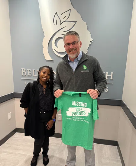 Man and woman standing indoors holding a green t-shirt with environmental message about missing 106+ pounds.