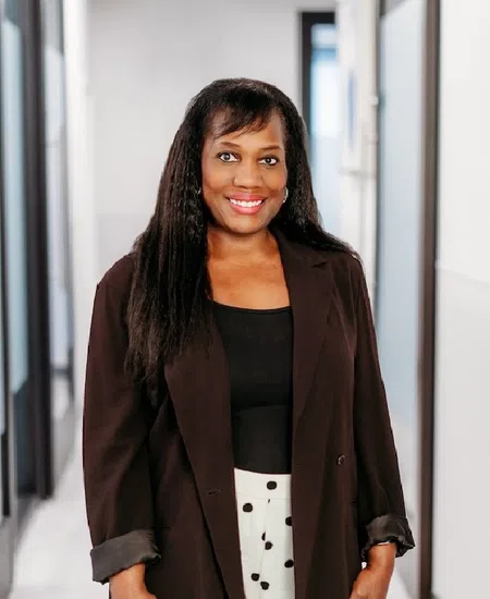 Smiling woman in black blazer and polka dot pants standing in a modern office hallway with glass doors.