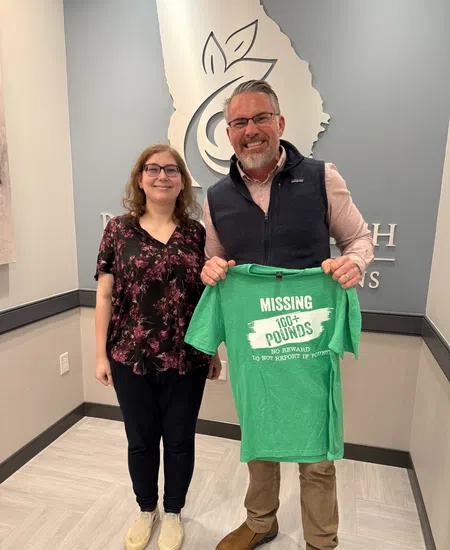 Man and woman smiling indoors holding a green shirt with weight loss message Missing 100 pounds to the happiness of fit.
