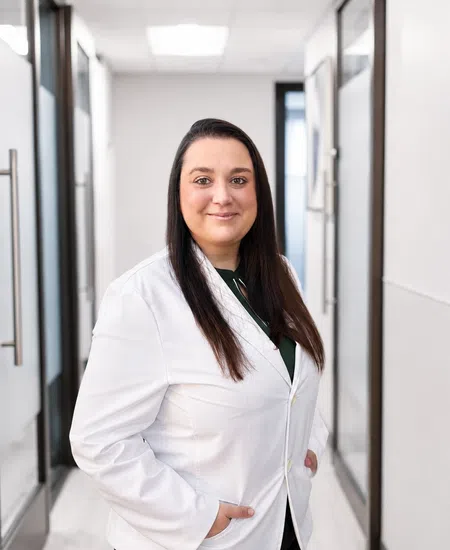 Confident female doctor wearing white coat standing in modern office hallway with hands in pockets.