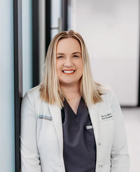 Smiling female nurse practitioner wearing white coat and dark scrubs with bright orange tie in medical hallway.
