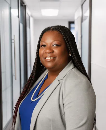 Smiling professional woman with braided hair wears gray blazer and blue top in office hallway.