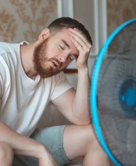 Tired man sitting in front of a fan, cooling off on a hot day indoors, wearing light clothes.