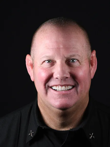 Smiling bald man in black shirt with crossed pins on collar against a black background