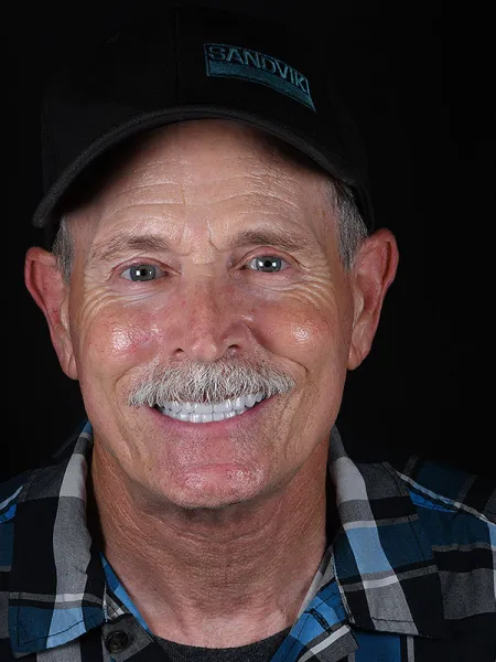 Smiling man wearing a black cap and plaid shirt against a dark background.