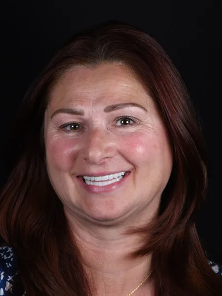 Smiling woman with long brown hair, wearing a gold necklace and blue patterned top, against a black background.