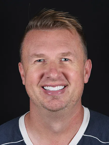 Smiling man with short blond hair wearing a navy blue shirt and white collar against a black background