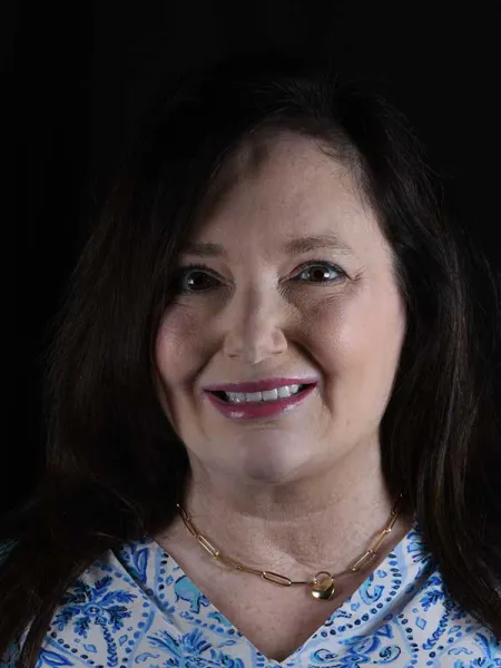 A smiling woman with long dark hair wearing a blue patterned blouse and a gold necklace against a black background.