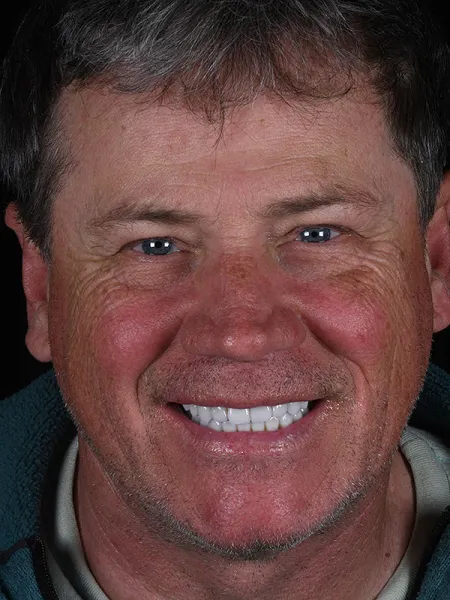 Close-up portrait of a smiling man with short hair and blue eyes against a dark background.