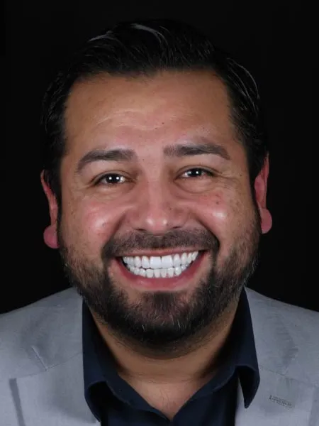 Smiling man with beard wearing a gray blazer and dark shirt against black background