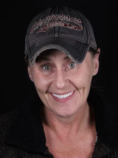 Smiling middle-aged woman wearing a weathered cap, against a dark background.