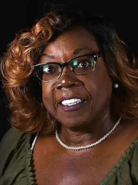 Smiling woman with curly brown hair, glasses, pearl necklace, and green blouse against black background.