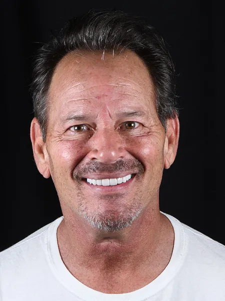 Smiling man with brown hair and a white T-shirt against a black background.