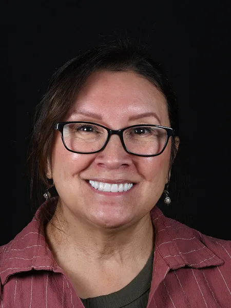 Smiling woman wearing glasses, with dark hair and a striped shirt, against a black background.
