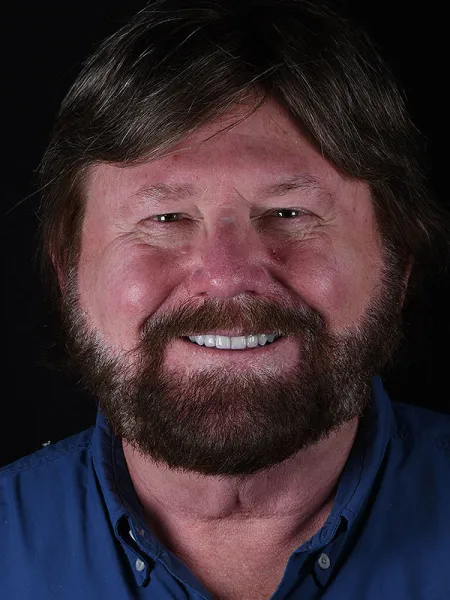 Smiling man with a beard wearing a blue shirt against a black background.