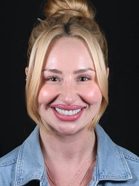 Smiling blonde woman with hair up wearing a denim jacket and layered necklaces against a black background