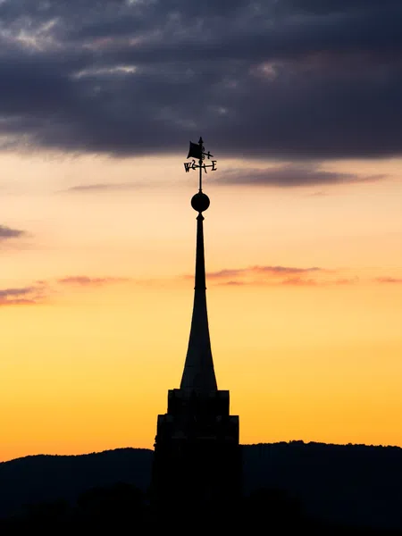 Silhouette of a pointed tower with a weather vane against a colorful sunset sky and distant hills.