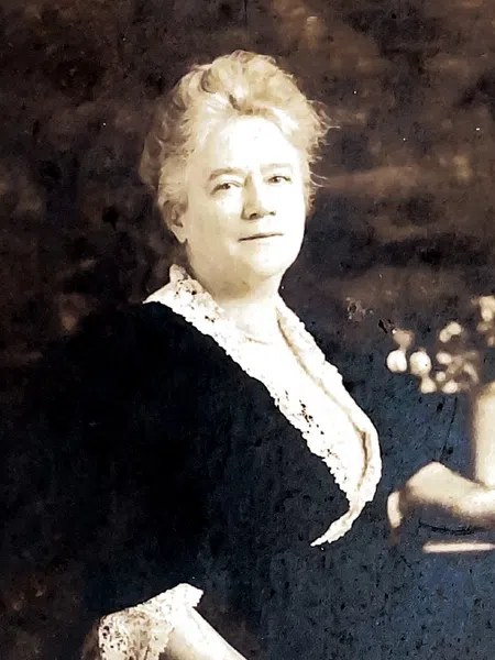 Vintage portrait of a woman in dark dress with lace collar and cuffs, posing beside a table with a vase of flowers.