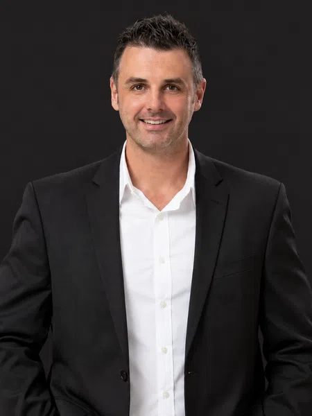 Smiling man in black blazer and white shirt posing confidently against a dark background