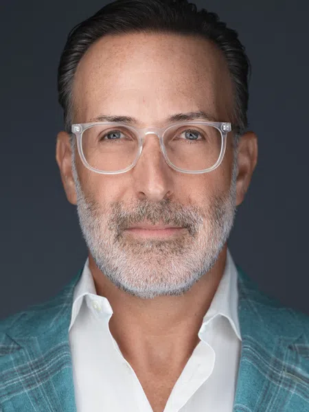 Middle-aged man with gray beard and clear glasses wearing a teal plaid jacket and white shirt against a dark background