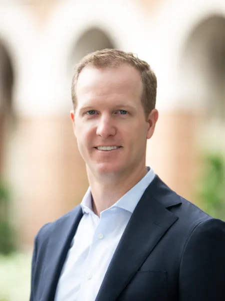 Professional man in blue suit and white shirt smiling in an outdoor setting with blurred background.