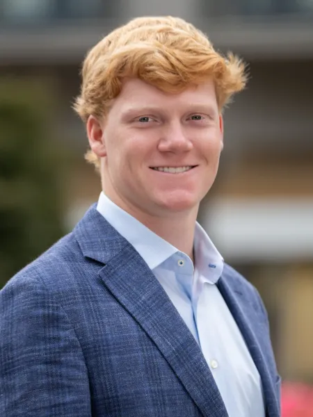 Young man with red hair wearing a blue suit jacket and white shirt smiling outdoors in daylight.