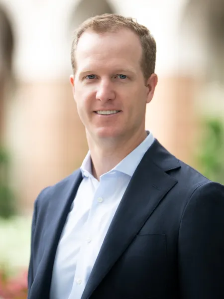 Professional man in blue suit and white shirt smiling in an outdoor setting with blurred background.